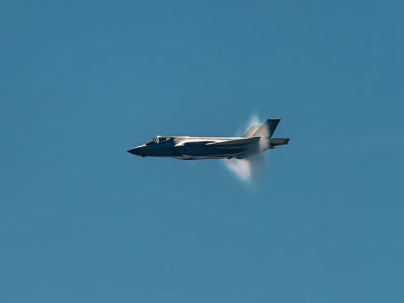 F-35A fighter jet in a high-speed pass with a vapour cone forming around its fuselage against a blue sky